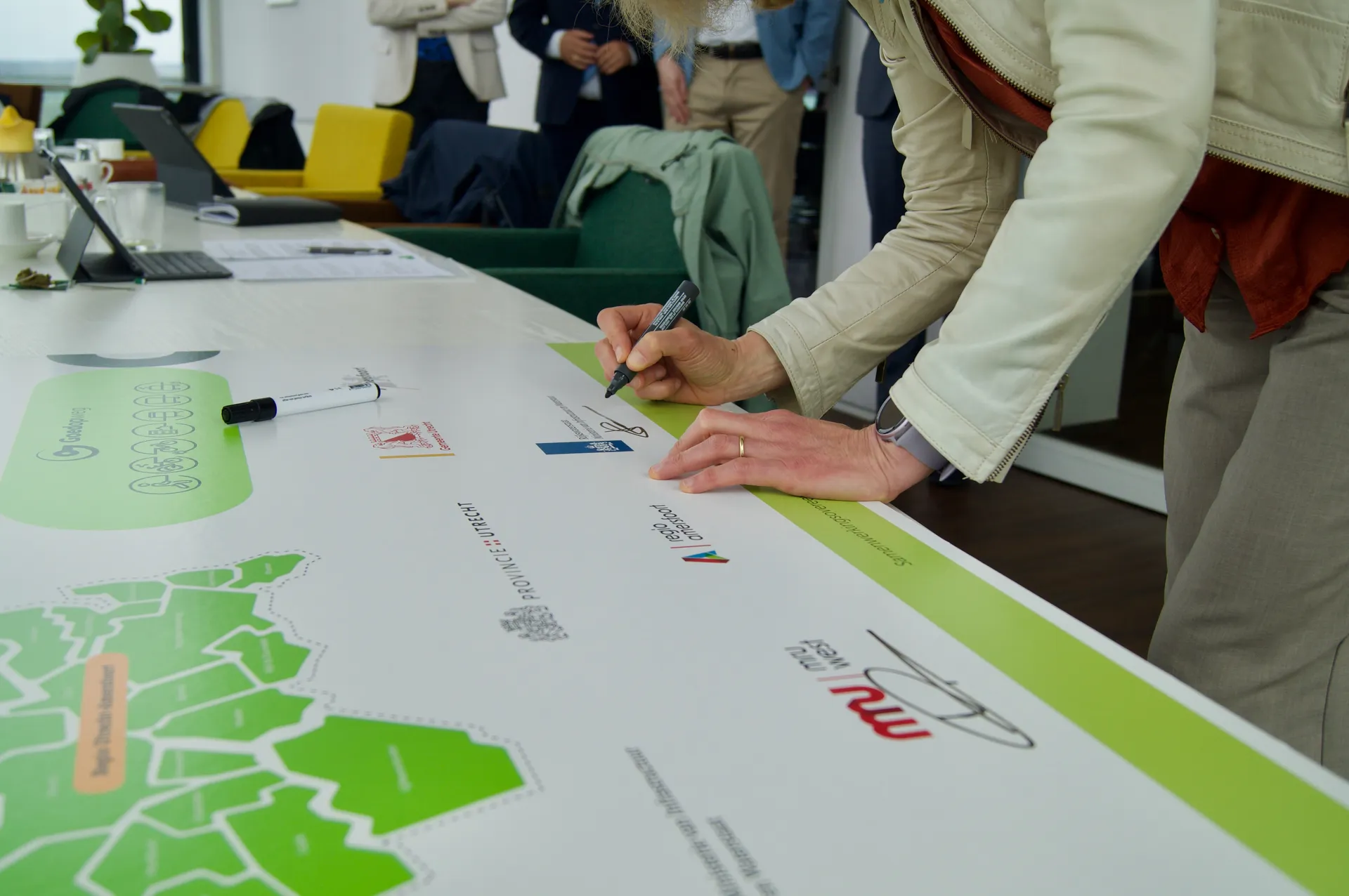 Kanselierondertekening op de grote ondertekeningsplaat in een vergaderruimte. Een volwassene tekent met een zwarte stift op een daarvoor gemarkeerde plek terwijl anderen toekijken, met een kaartachtige illustratie en diverse logo’s op de plaat.