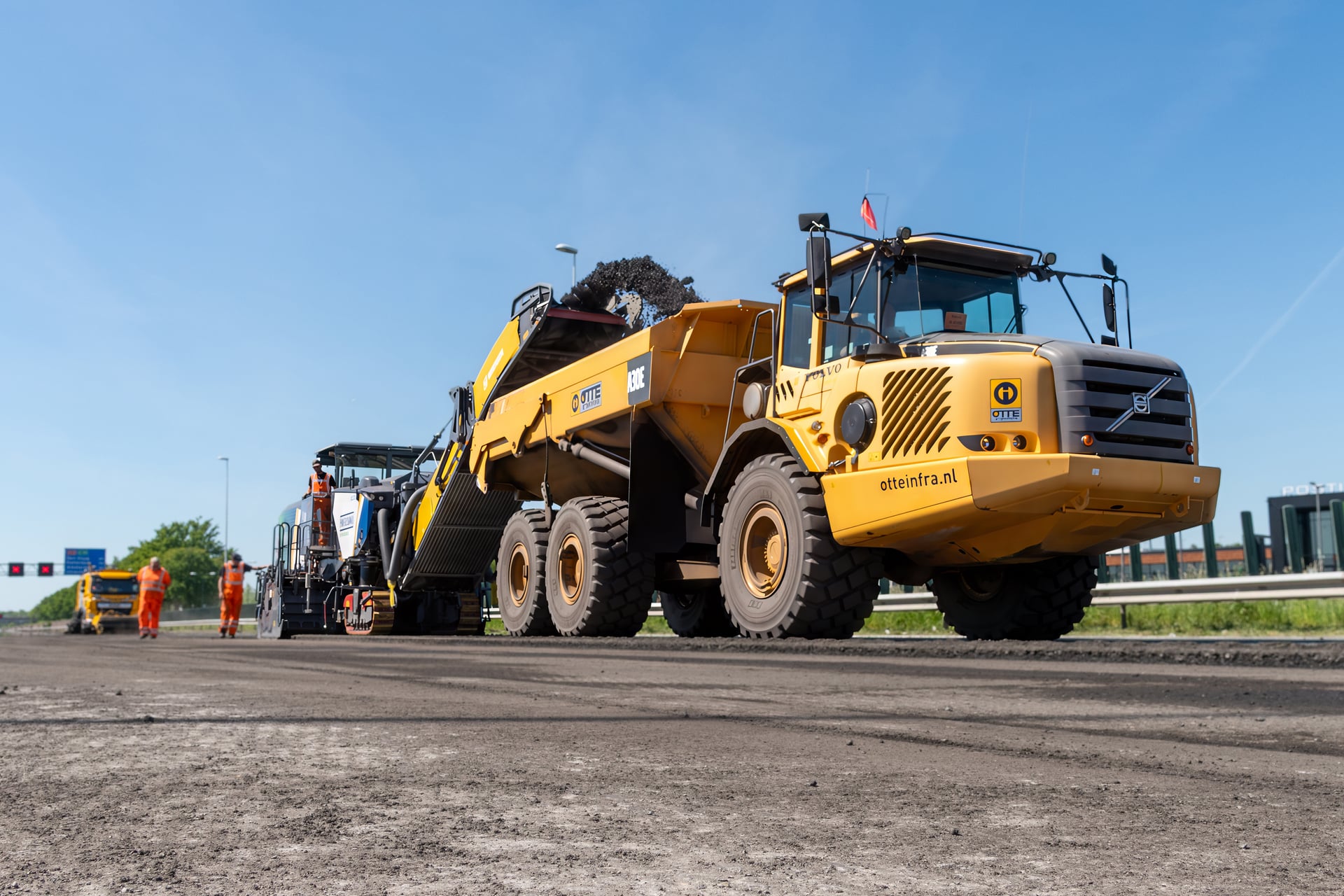 OTTE, een grote gele Volvo-kieptruck met knikgestuurd chassis, kantelt zijn bak om asfalt in de trechter van een asfaltspreidmachine te laten storten tijdens grootschalige wegwerkzaamheden; links op de afgesloten rijbaan werken wegwerkers in feloranje veiligheidskleding en is er portaalkunst/afzetting zichtbaar.