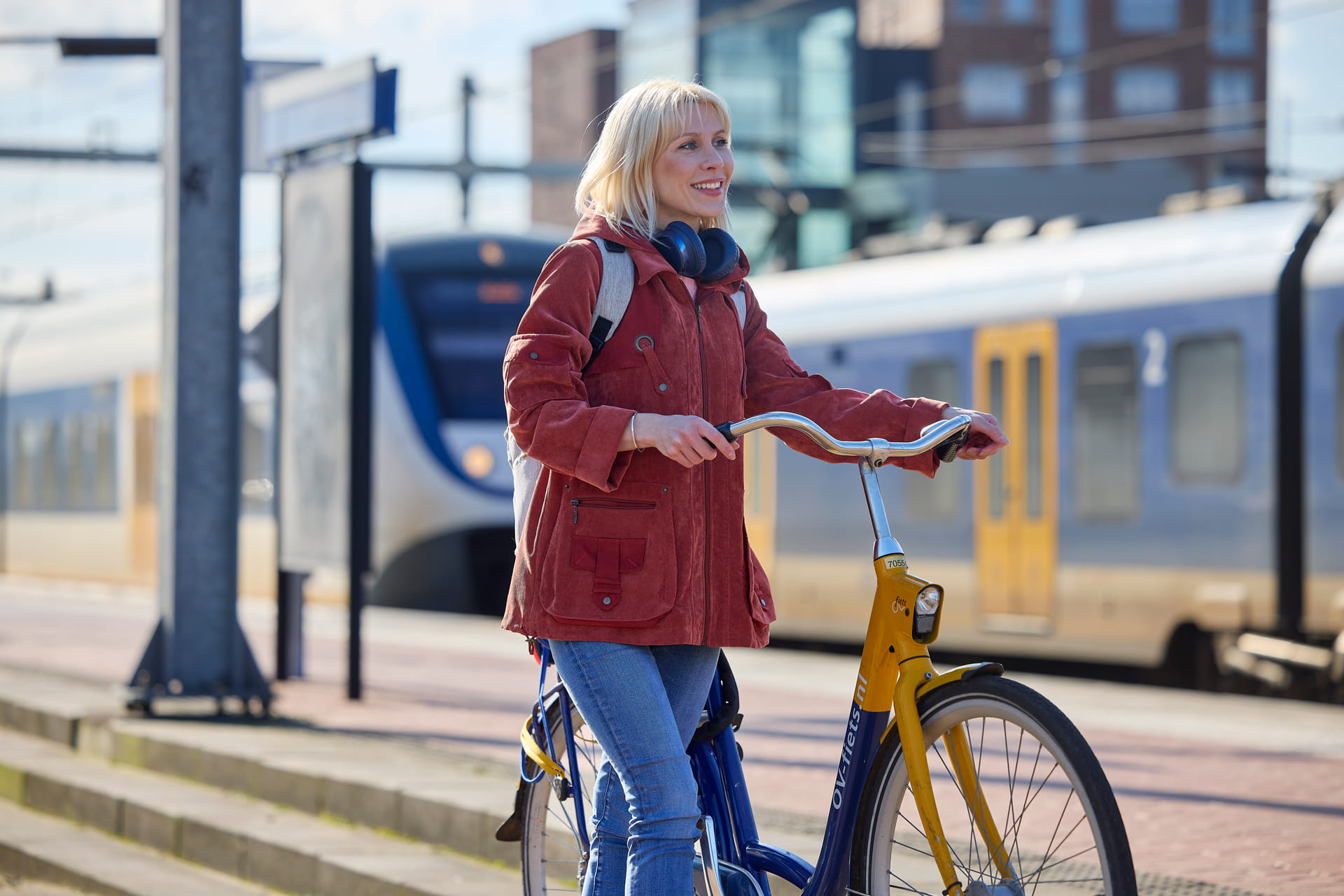 Een dame die naast haar fiets staat. Op de achtergrond staan er twee treinen stil.