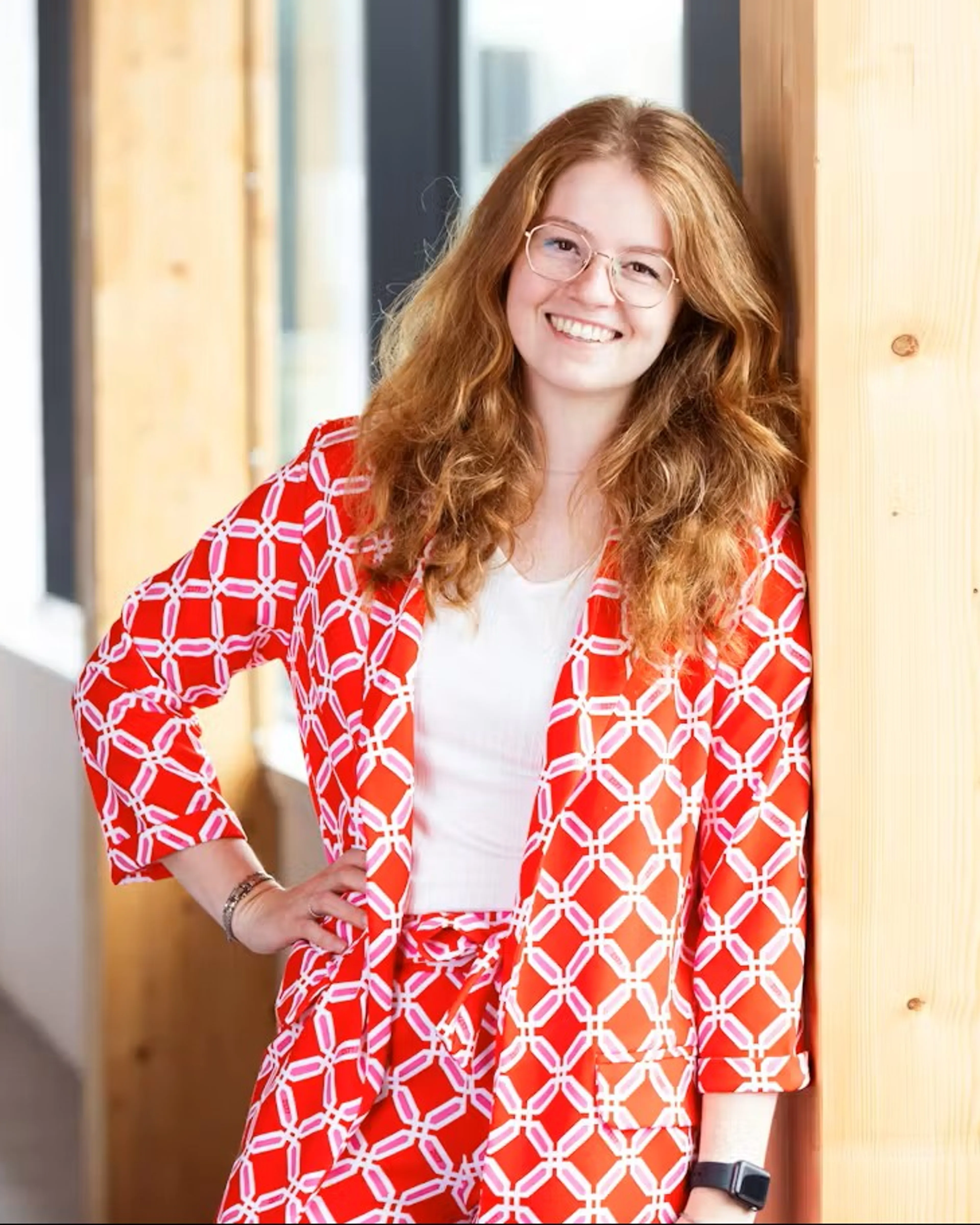 Model beschrijft een jonge vrouw met lang, golvend rood haar en een bril, glimlachend tegen een brede houten kolom leunend in een lichte, moderne gang met grote ramen aan de rechterkant. Ze draagt een opvallend rood tweedelig pak met een wit geometrisch patroon over een wit shirt, één hand op de heup en een horloge zichtbaar op de pols; de omgeving heeft warme houten accenten en veel daglicht, terwijl de achtergrond zacht onscherp blijft en de nadruk ligt op haar ontspannen, zelfverzekerde pose.