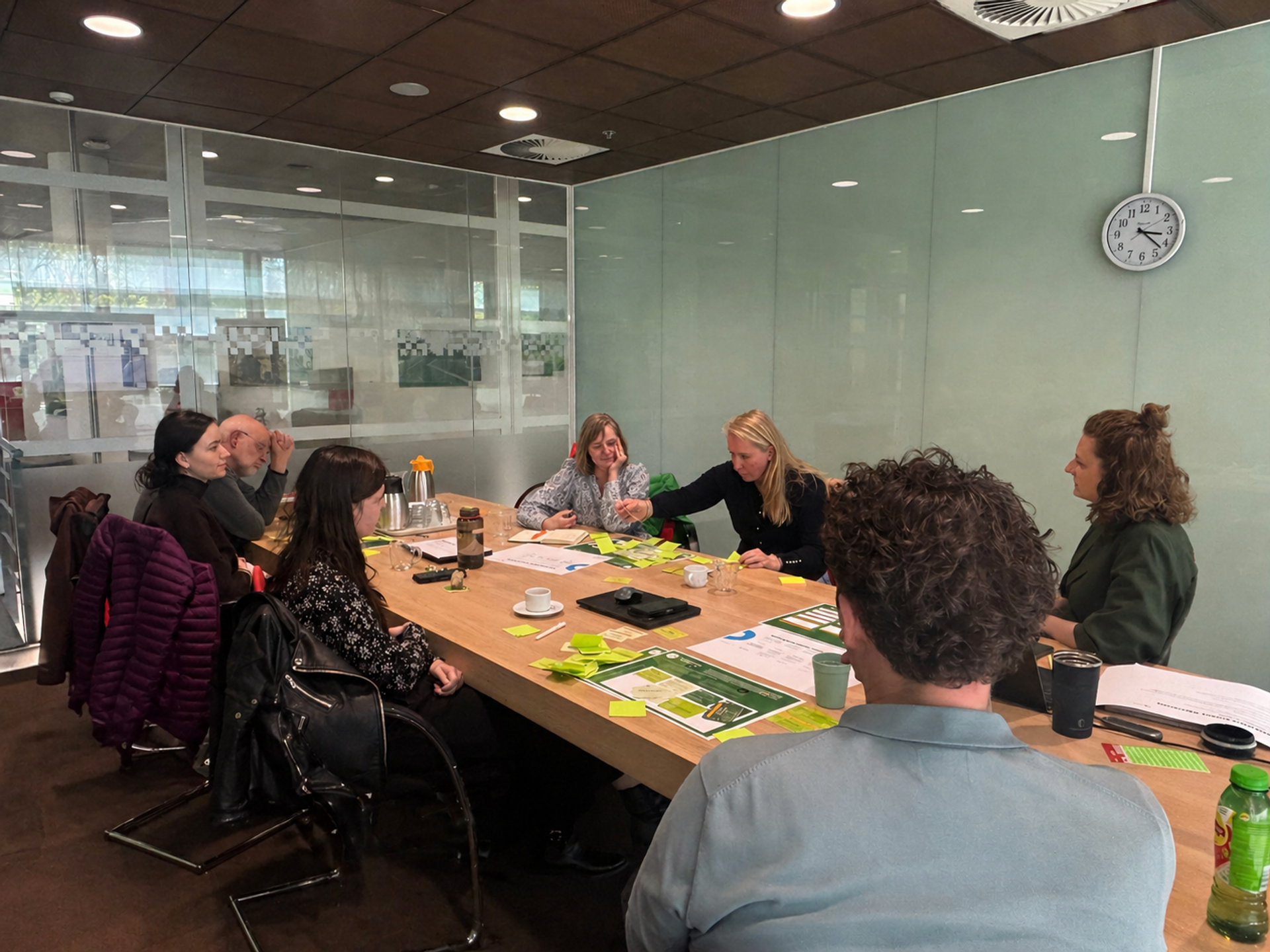 Vergaderruimtebijeenkomst: Een groep van ongeveer zeven volwassenen zit rondom een lange houten tafel in een kantooromgeving, waar geprinte werkbladen, post-its, pennen en drankjes aanwezig zijn terwijl een blonde vrouw voorover leunt en wijst naar notities en post-its. De setting duidt op een geconcentreerde werksessie of brainstorm.