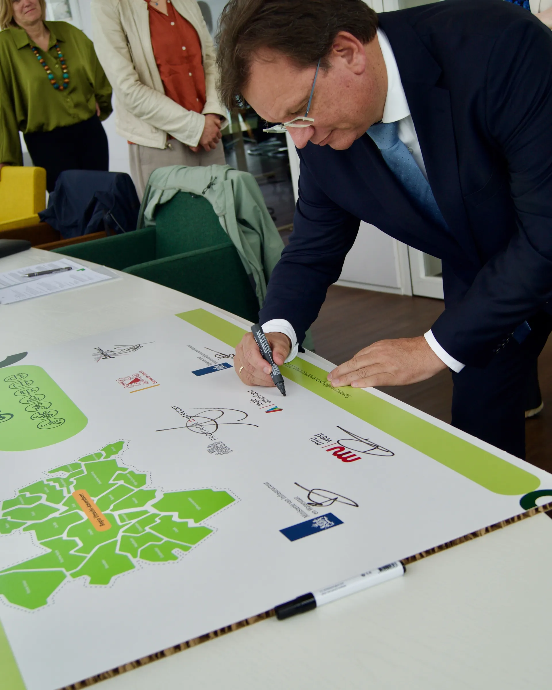 Kleine ondertekeningsmoment in een kantooromgeving. Een middelbare man in een donkerblauw pak zet met een dikke zwarte stift zijn handtekening op een liggende presentatieplaat met een groene rand en verschillende logo’s, terwijl twee volwassenen toekijken op de achtergrond.