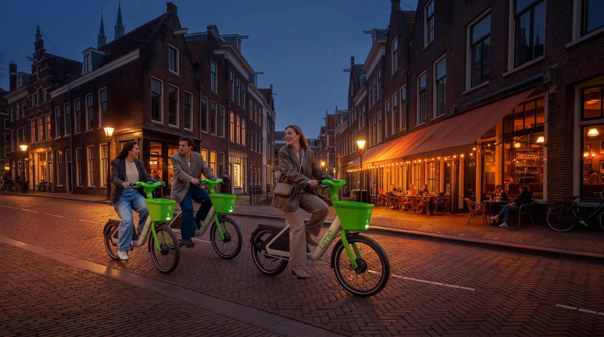 Drie Lime-deelfietsen rijden door een sfeervolle historische binnenstad op een met klinkers bestrate straat; twee fietsers links lachen naar elkaar, terwijl de voorste vrouw vooruit fietst in een beige/taupe outfit. De straat wordt warm verlicht door straatlantaarns en de ramen van smalle panden, en aan de rechterkant is een café met oranje luifel en tafeltjes gevuld met gasten, terwijl kerk- of torenspitsen tegen een donkerblauwe schemerlucht op de achtergrond te zien zijn.