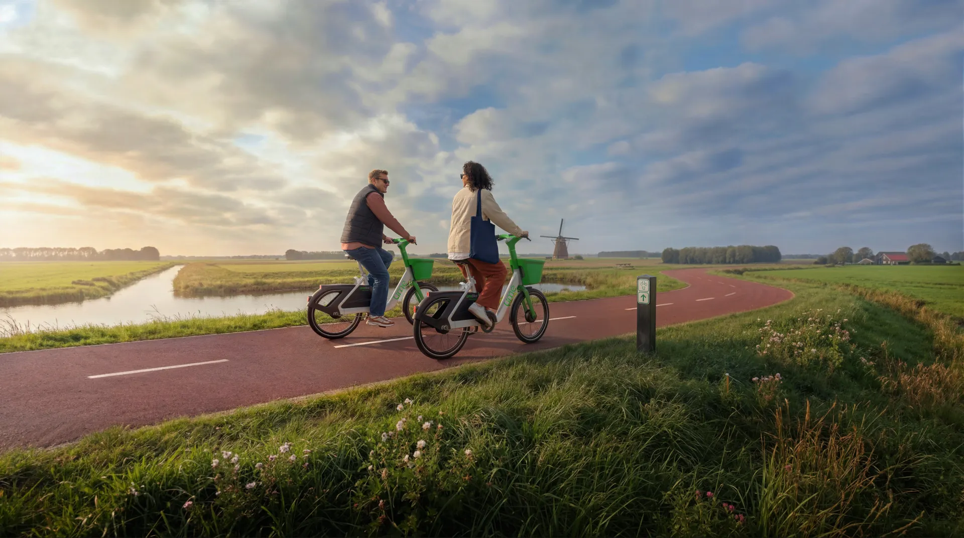Op een rood geasfalteerd fietspad in een uitgestrekt polderlandschap fietsen twee volwassenen naast elkaar op felgroene fietsen, terwijl ze elkaar aankijken alsof ze in gesprek zijn. Op de voorgrond staat een klein groen-wit routepaaltje langs de berm met wilde bloemen en gras, en op de horizon zijn bomen, boerderijen en een traditionele windmolen zichtbaar tegen een lucht gevuld met brede wolkenpartijen in warm, laag zonlicht.