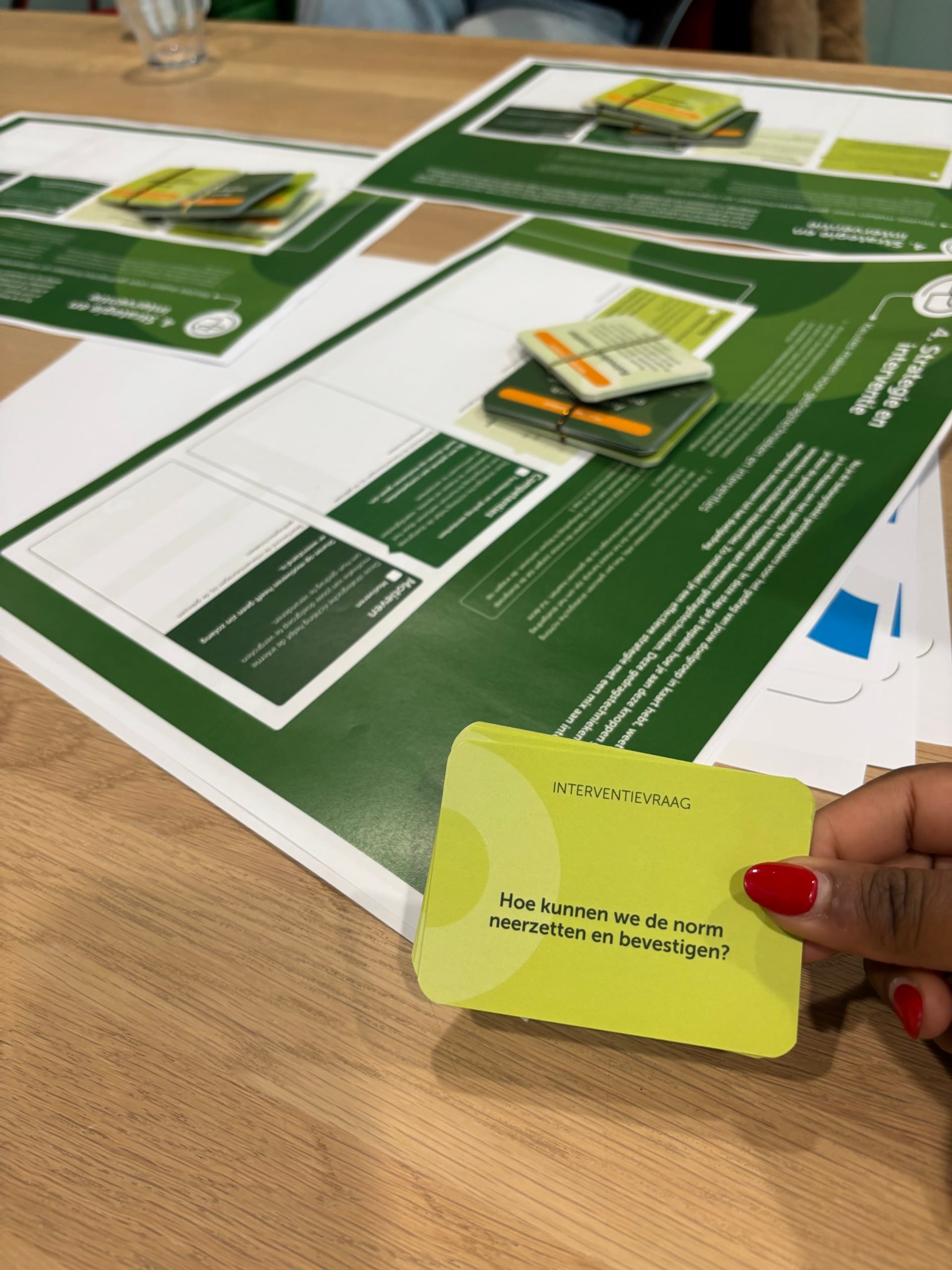 Interventievraag: Hoe kunnen we de norm neerzetten en bevestigen? Een houten tafel met groen-witte werkbladen voor een workshop, waarop meerdere kaarten met elastiekjes bijeen gehouden zijn; op de voorgrond houdt een hand met felrode nagellak een lichtgroene kaart omhoog.
