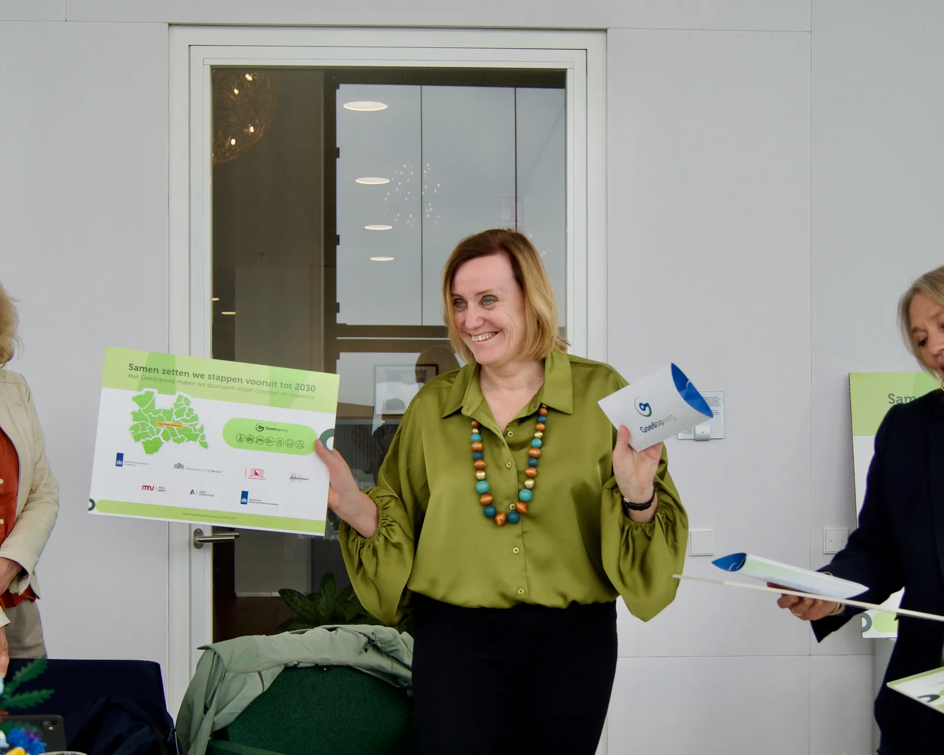 Nederland – In een lichte, moderne vergaderruimte staat een glimlachende middelbare vrouw centraal, met halflang blond haar en een olijfgroene blouse, terwijl ze presentatiemateriaal omhoog houdt en een grote groen-witte poster met tekst “Samen zetten we stappen vooruit tot 2030” en “Groningen” draagt. Ze lijkt een formeel moment toe te lichten of vieren, mogelijk tijdens een presentatie.