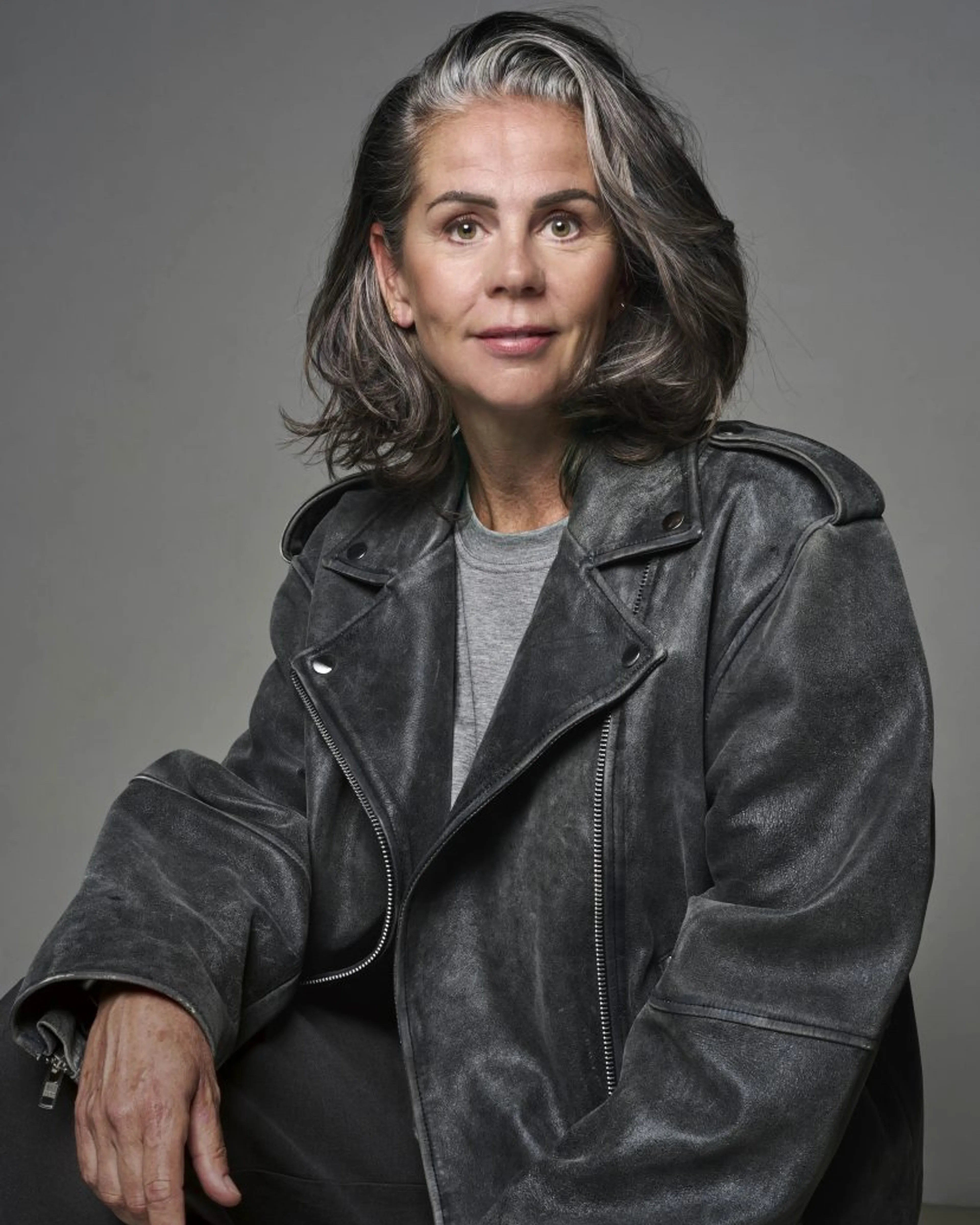 Er is één vrouwelijk portret op een egale grijze achtergrond. Het beeld toont een middelbare vrouw in een donkere leren bikerjack en grijs T‑shirt, zittend met een ontspannen houding en een kalme, directe blik naar de camera.