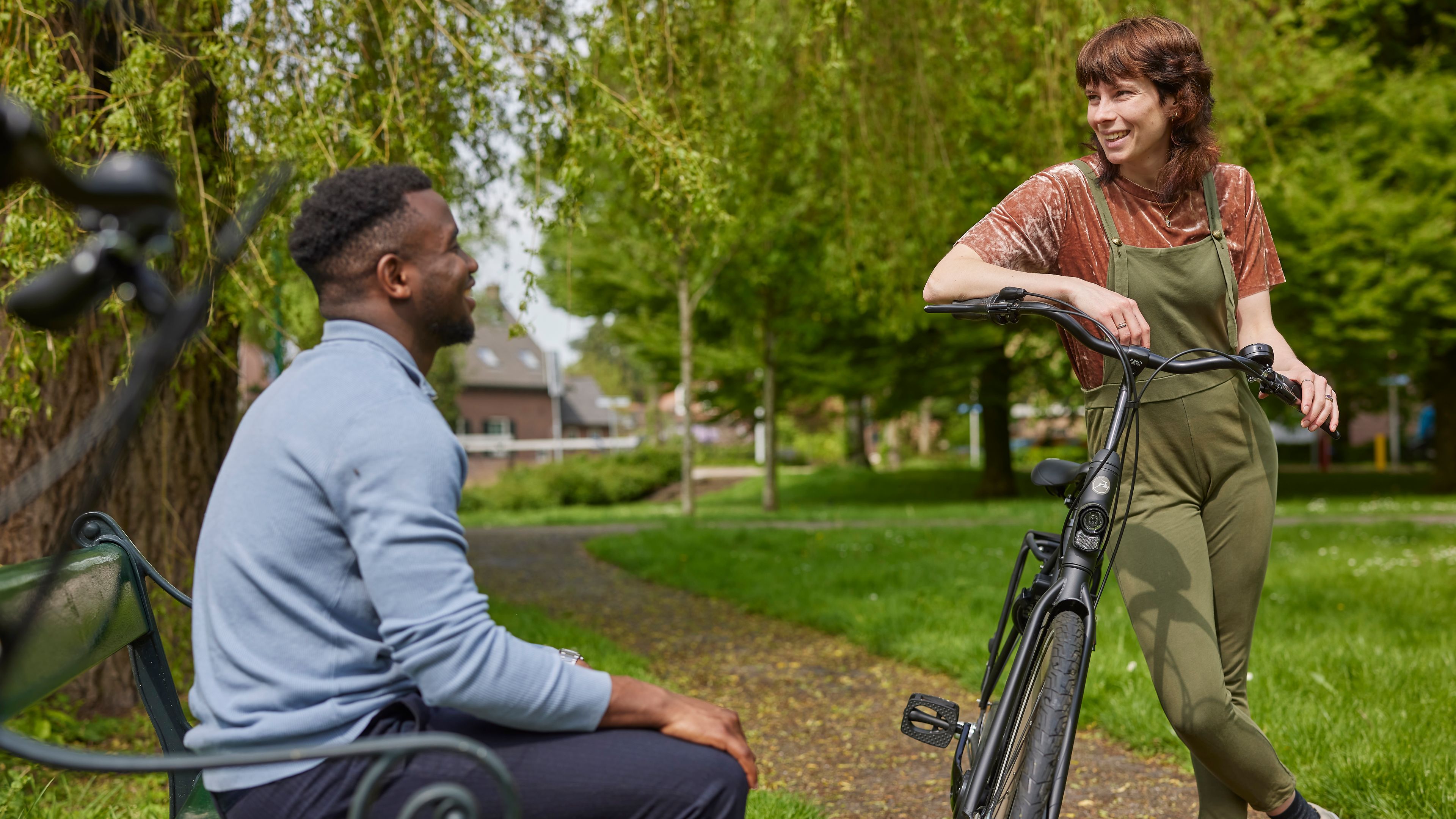 Man zit op een bankje in het park. Hij praat met een vrouw die tegen haar fiets aan leunt.