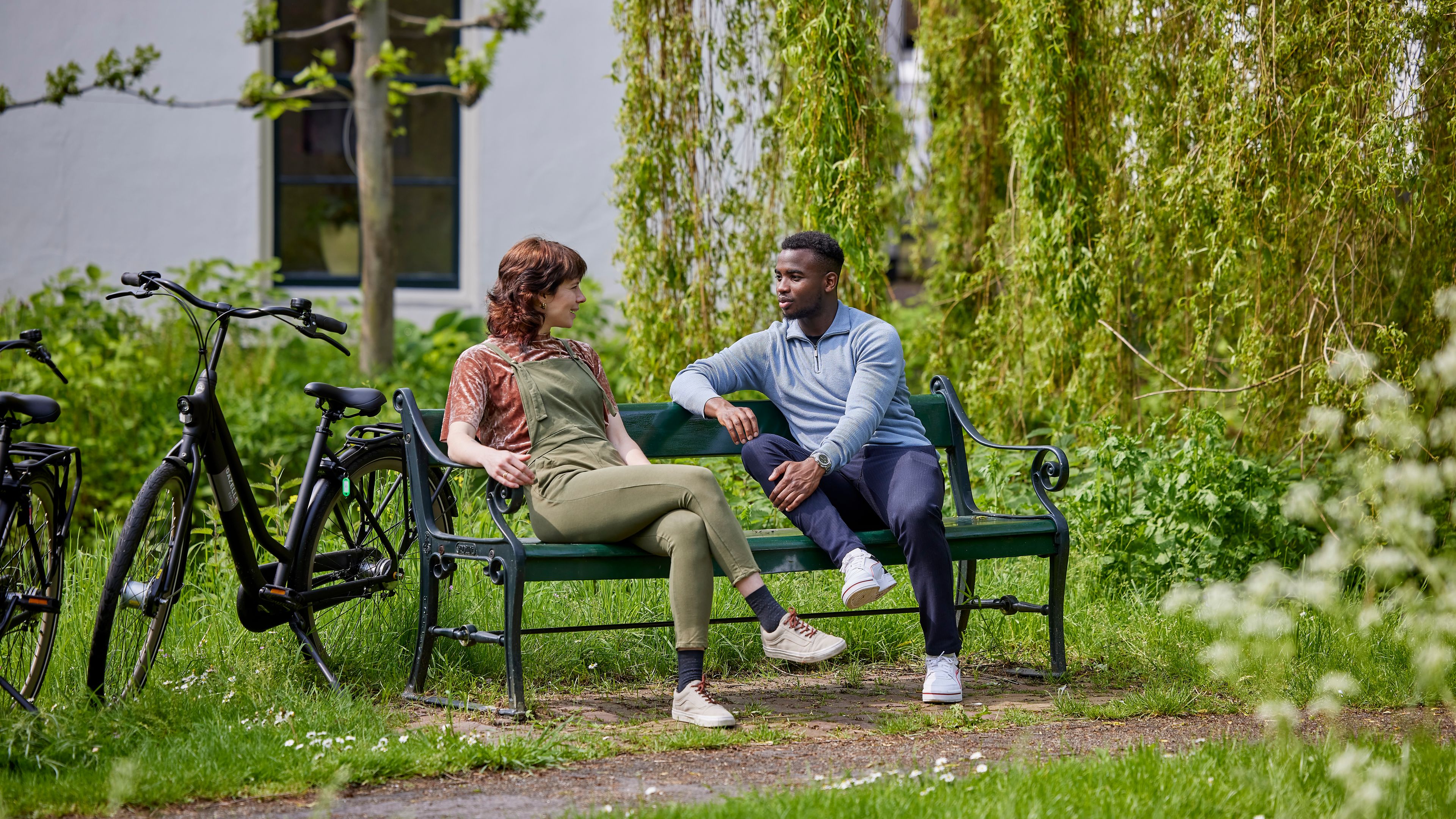 Man en vrouw zitten te praten op een bankje in het park. Naast hen staan twee fietsen.