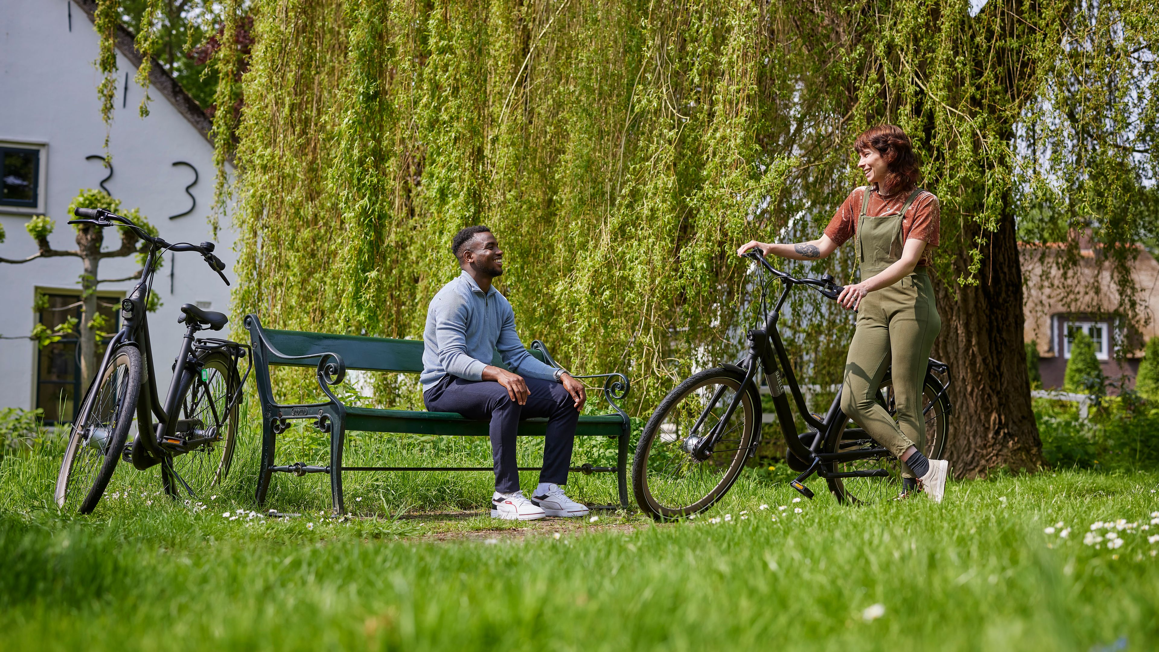 Man zit op een bankje in het park. Naast hem staat zijn fiets. Hij praat met een vrouw die tegen haar fiets aan leunt.