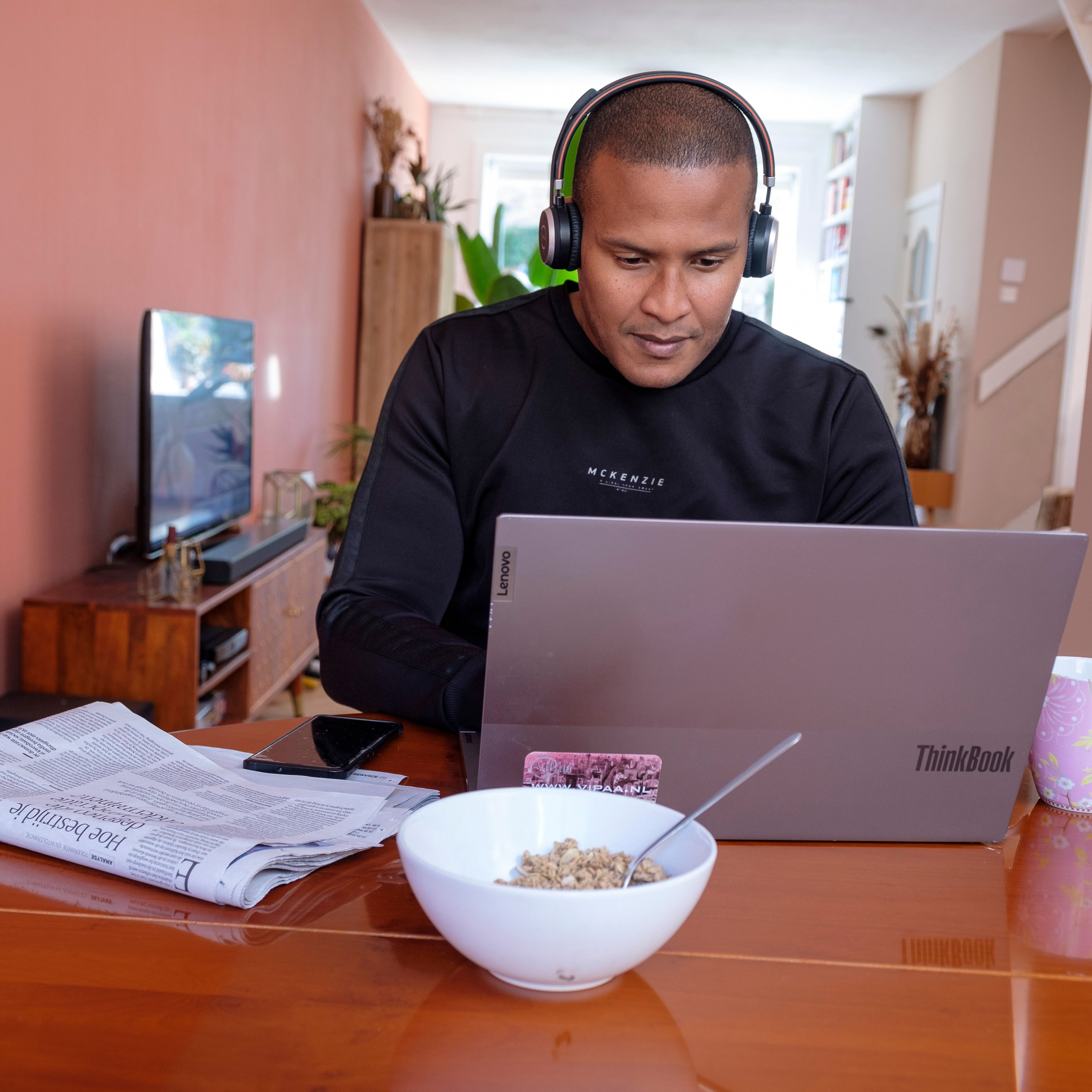 Een man zit met een koptelefoon aan tafel thuis achter zijn laptop.