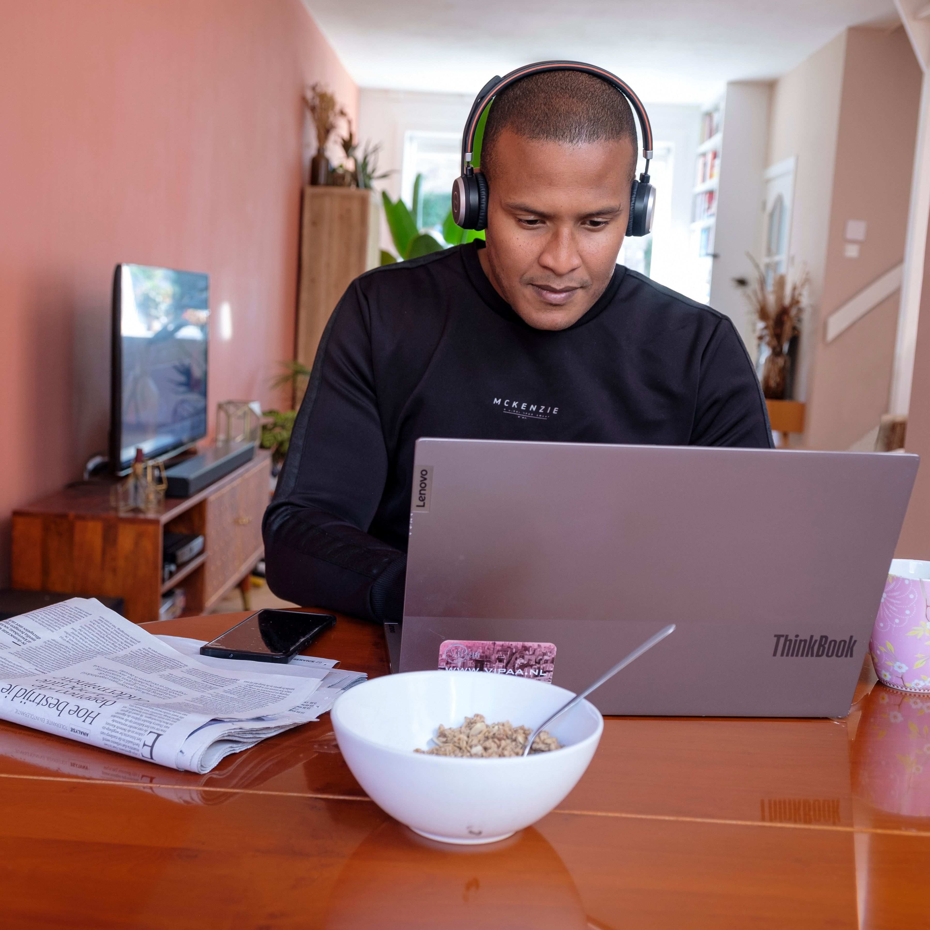 Een man zit met een koptelefoon aan tafel thuis achter zijn laptop.