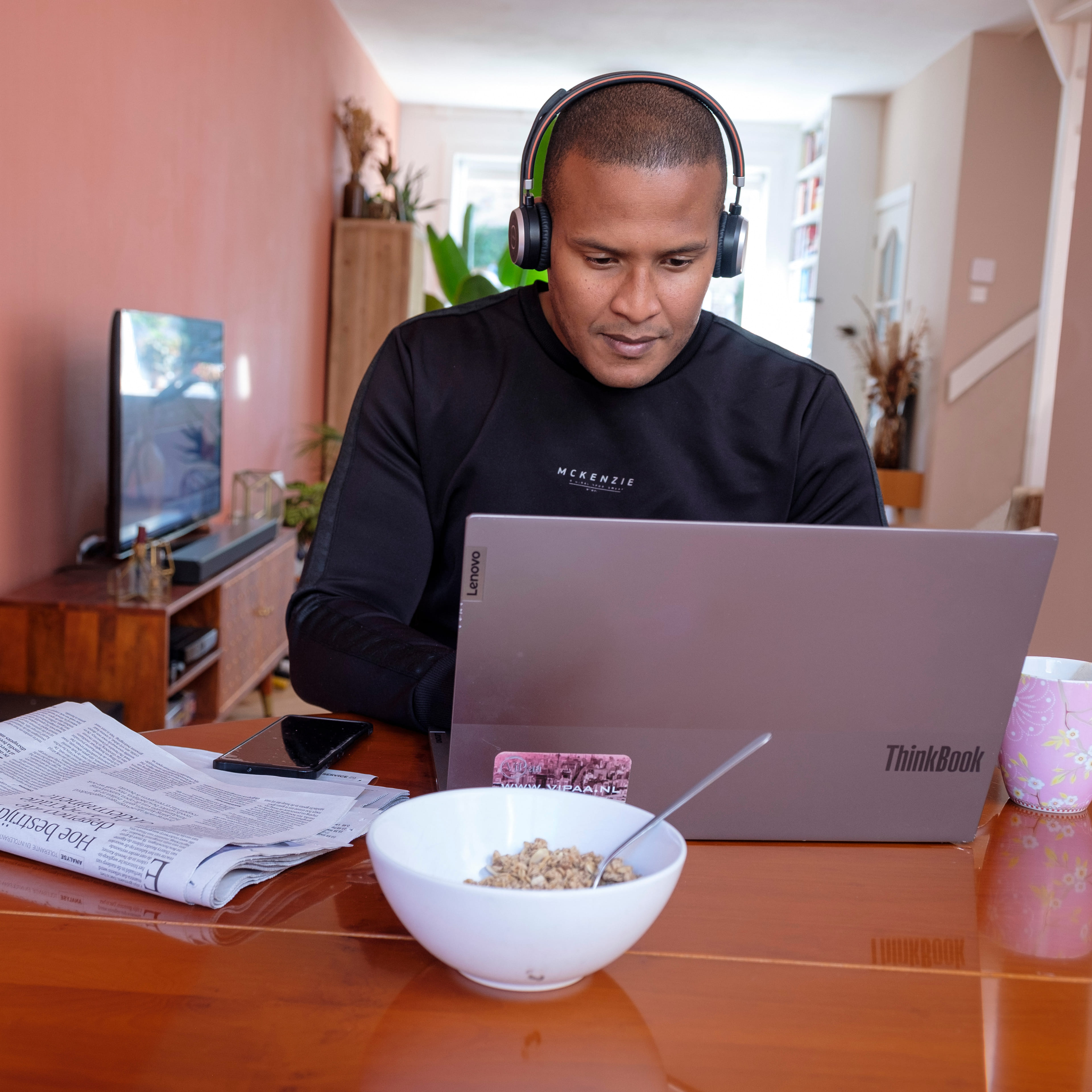 Een man zit met een koptelefoon aan tafel thuis achter zijn laptop.