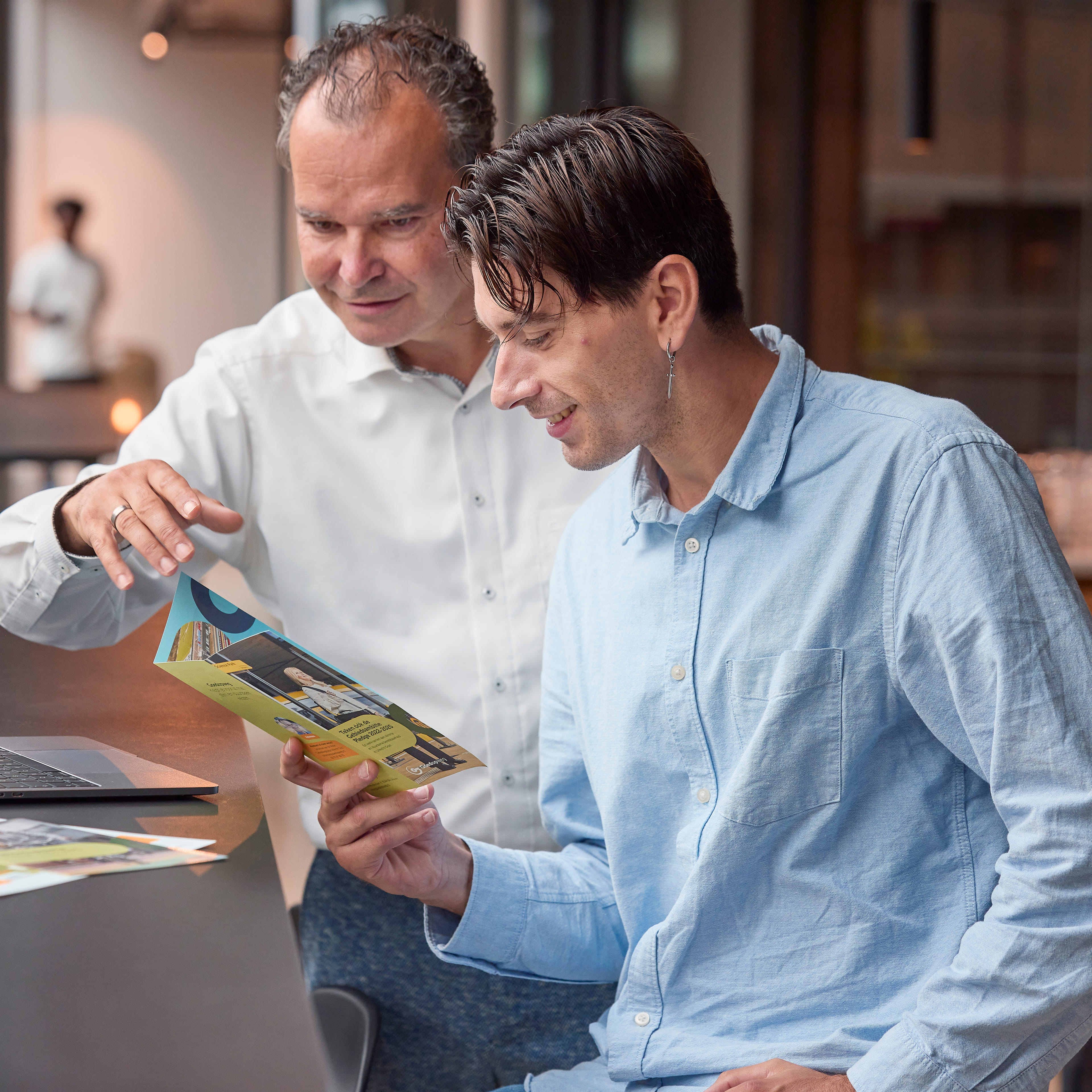 Twee collega's met elkaar in gesprek. Ze kijken samen naar een flyer.