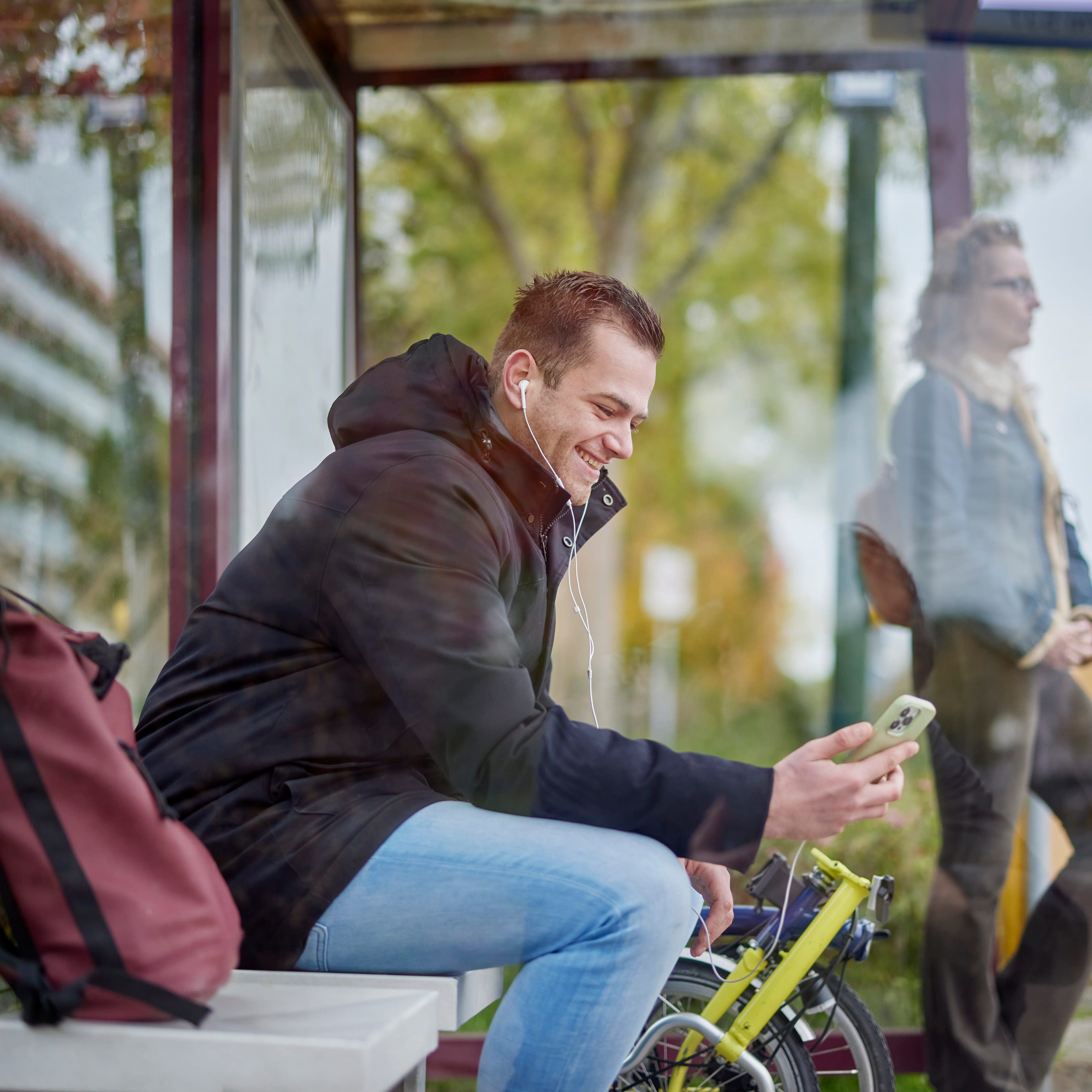 Jonge man zit in een bushalte met zijn telefoon in zijn hand. Naast hem staat een vouwfiets