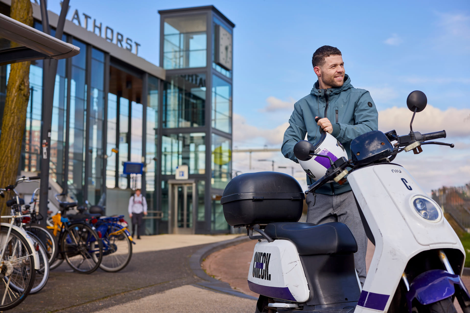 Je ziet een man met in zijn handen een helm van een check deelscooter. De check scooter staat naast hem. Hij staat voor station Amersfoort Vathorst