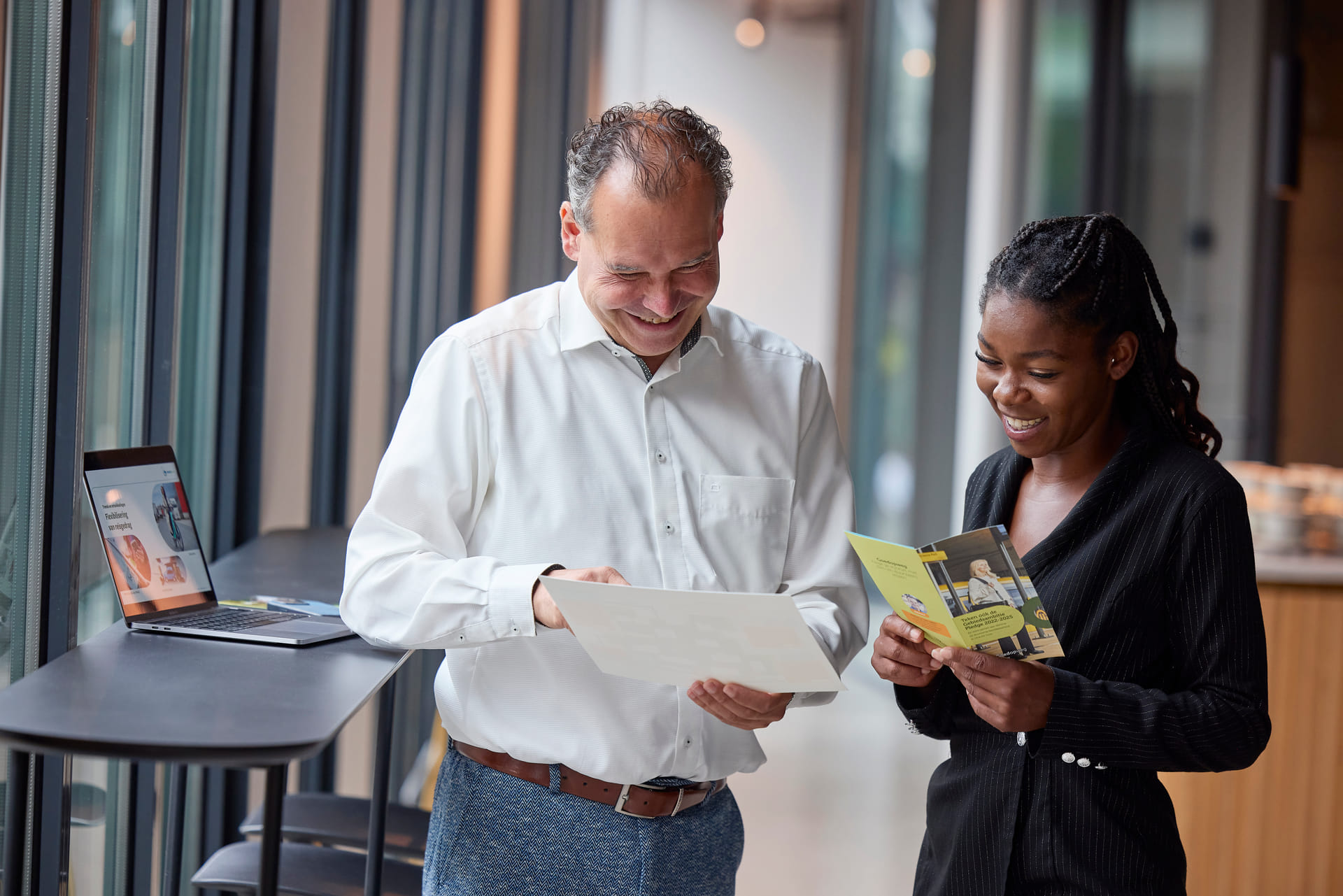 Een mannelijke en vrouwelijke collega met elkaar in gesprek. Ze kijken samen naar een flyer.