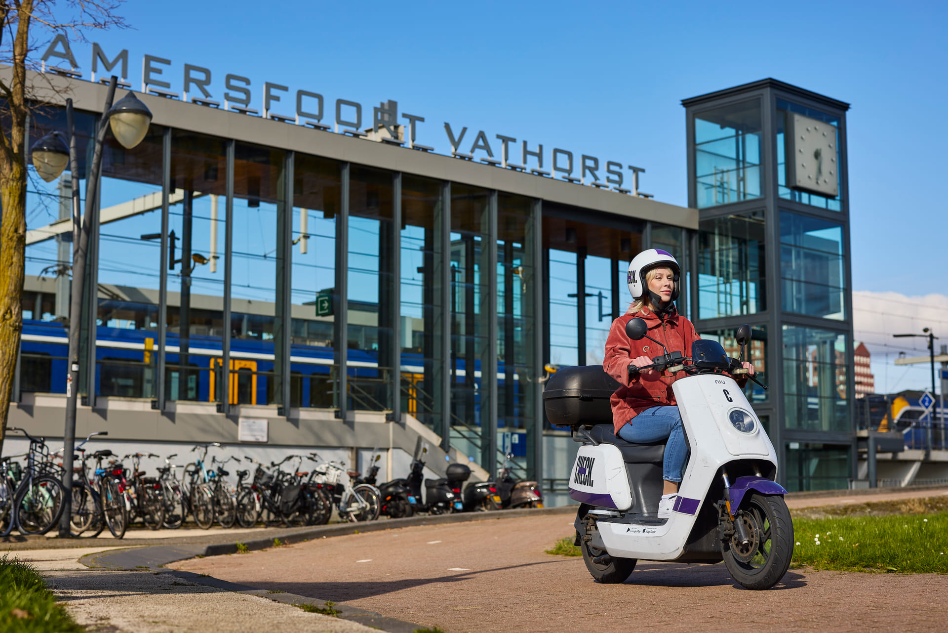 Je ziet een vrouw op een check deelscooter zitten. Ze staat voor station Amersfoort Vathorst