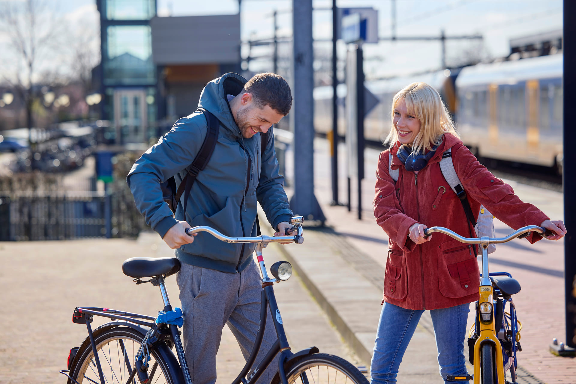 Je ziet een man en vrouw met elkaar lachen. Ze hebben beiden een fiets vast. Ze staan op station Amersfoort Vathorst.