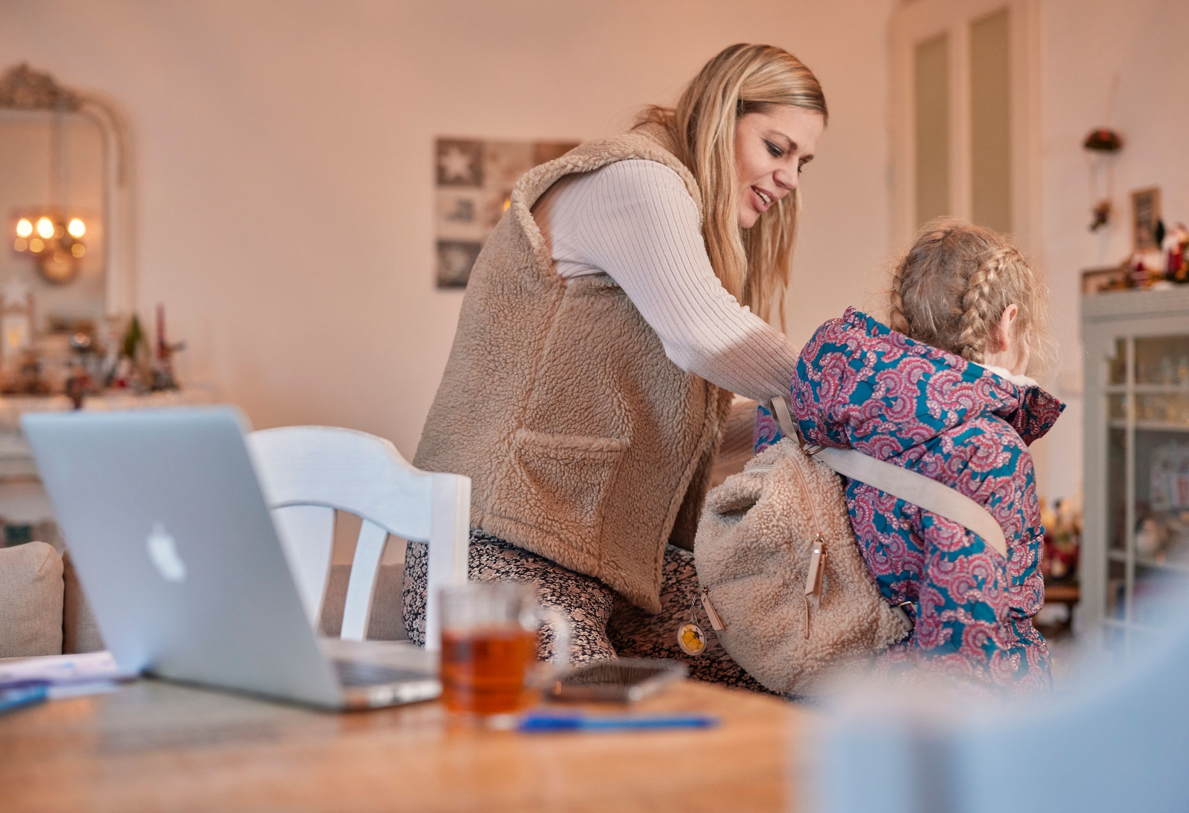 Een vrouw helpt een kind met haar jas en rugtas. Op tafel staat een opengeklapte laptop en een kop thee.