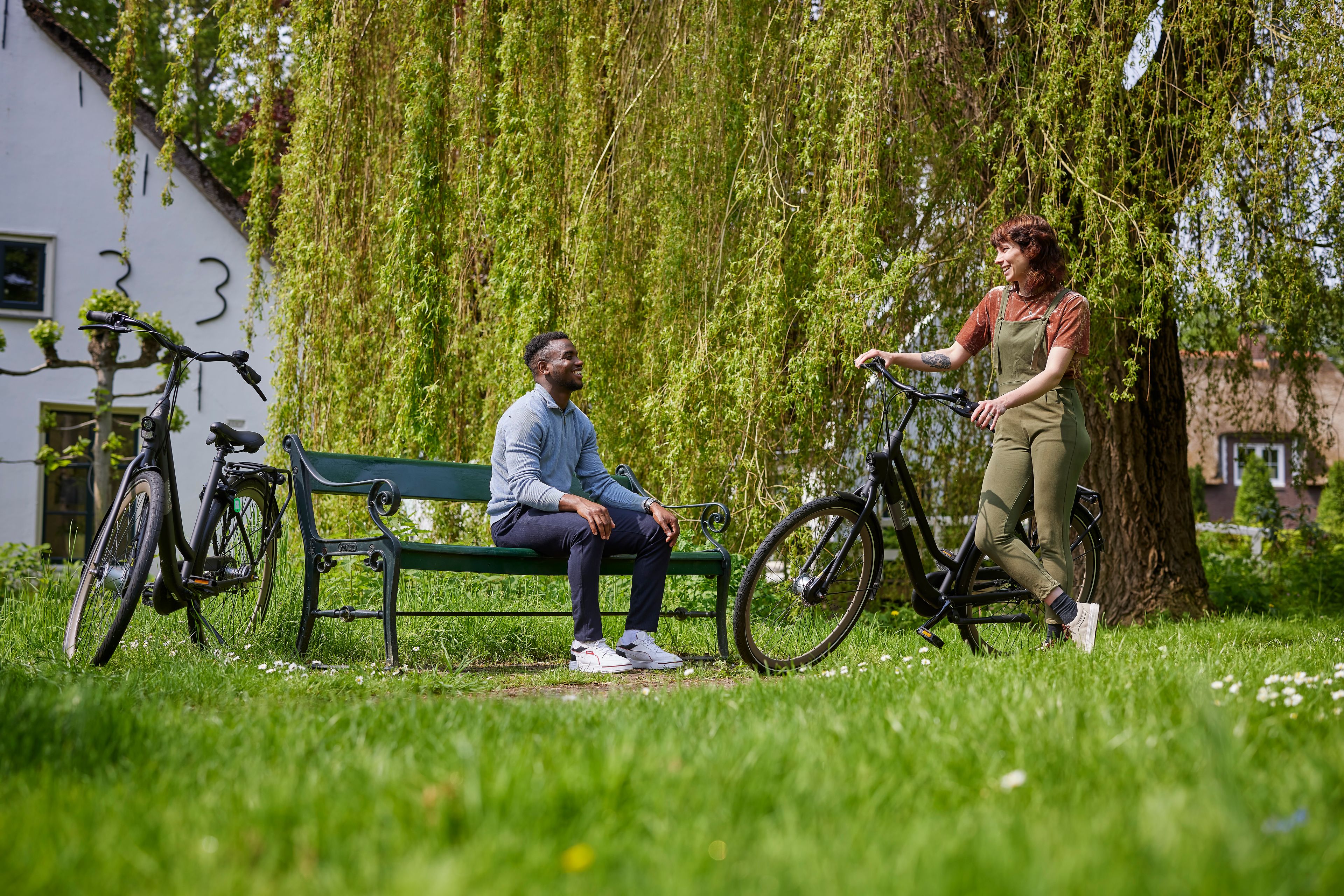 Man zit op een bankje in het park. Naast hem staat zijn fiets. Hij praat met een vrouw die tegen haar fiets aan leunt.