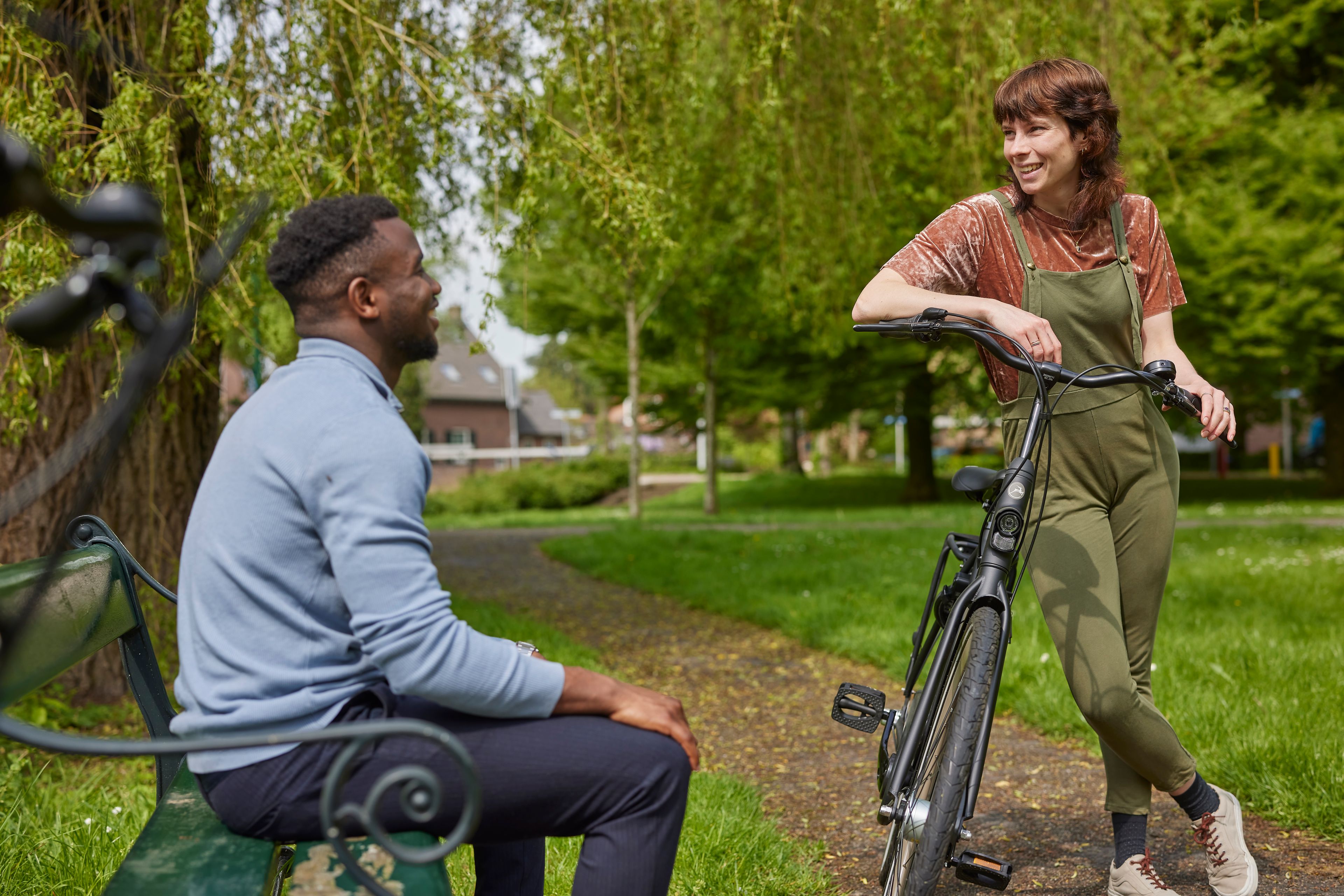 Man zit op een bankje in het park. Hij praat met een vrouw die tegen haar fiets aan leunt.