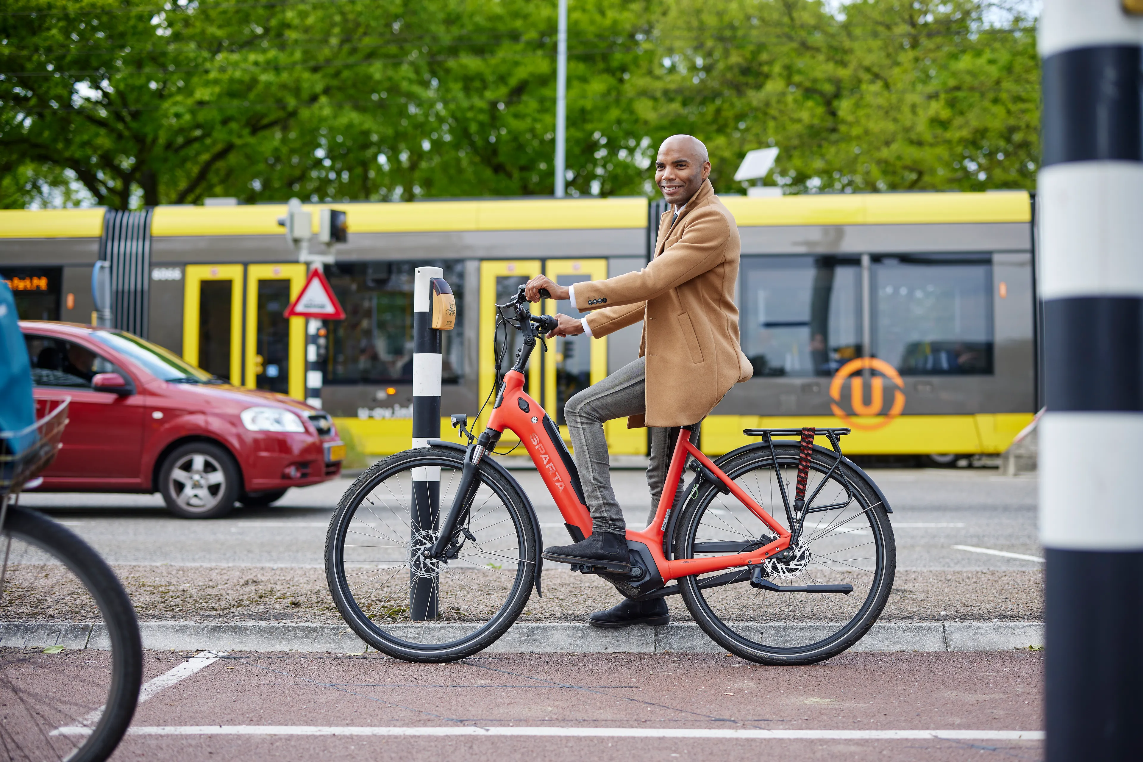Man staat op een deelfiets te wachten bij het stoplicht. Achter hem rijdt een tram.